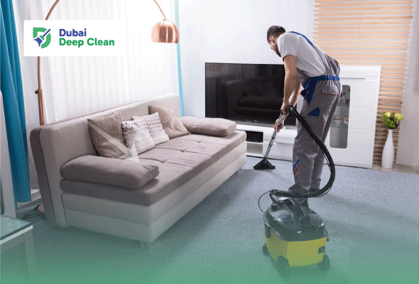 A professional cleaner in a grey and blue uniform using an industrial vacuum cleaner to deep clean the carpeted floor of a modern villa living room near a grey sofa.