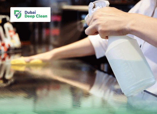 A close-up of a cleaning professional using a spray bottle and a yellow microfiber cloth to sanitize a reflective high-gloss countertop in a commercial club or lounge area.