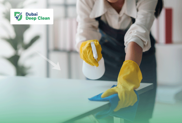 A cleaning professional wearing yellow safety gloves using a disinfectant spray and blue microfiber cloth to sanitize a white office desk.